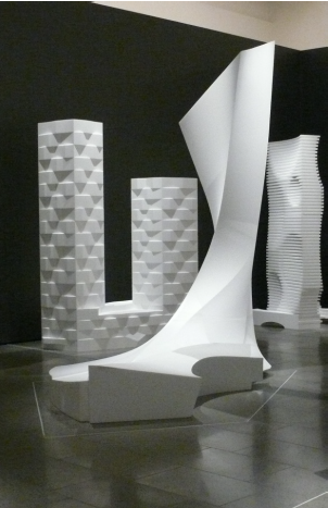 White parametric architectural models — faceted towers and a twisting form — in a gallery